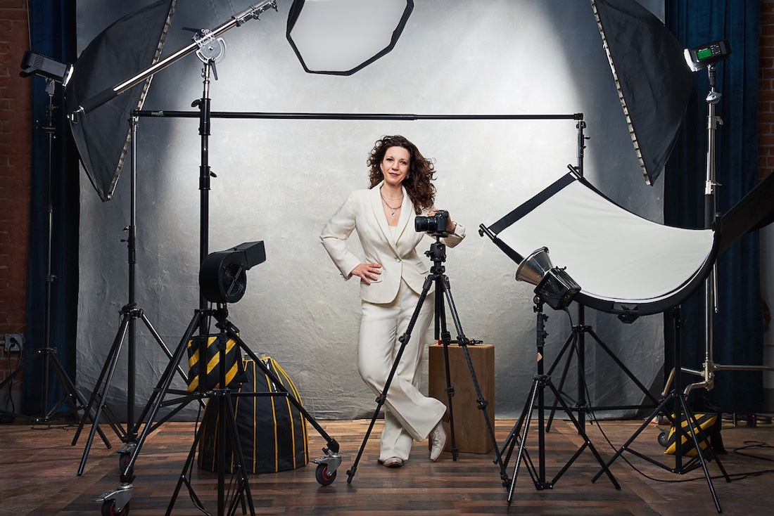 A woman in a white suit poses confidently in a professional photography studio. She is surrounded by cameras, lights, and studio equipment, standing against a gray backdrop on a wooden floor.