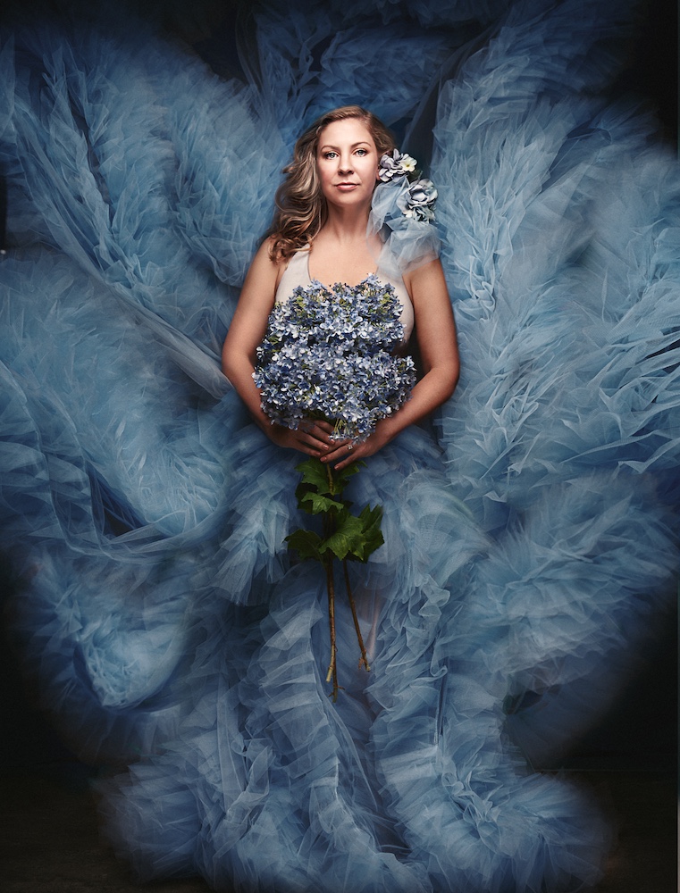 A woman stands against a dark backdrop, wearing an elaborate blue tulle gown that fans out around her. She holds a bouquet of blue flowers, with a matching flower in her hair, creating a serene and ethereal atmosphere.