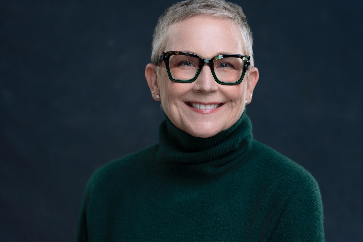 A woman with short gray hair and glasses smiles warmly. She is wearing a dark green turtleneck sweater against a dark, plain background.