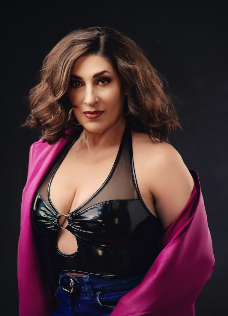 A person with wavy brown hair poses confidently against a dark background, wearing a black halter top with a ring detail and a deep magenta jacket draped over their shoulders.