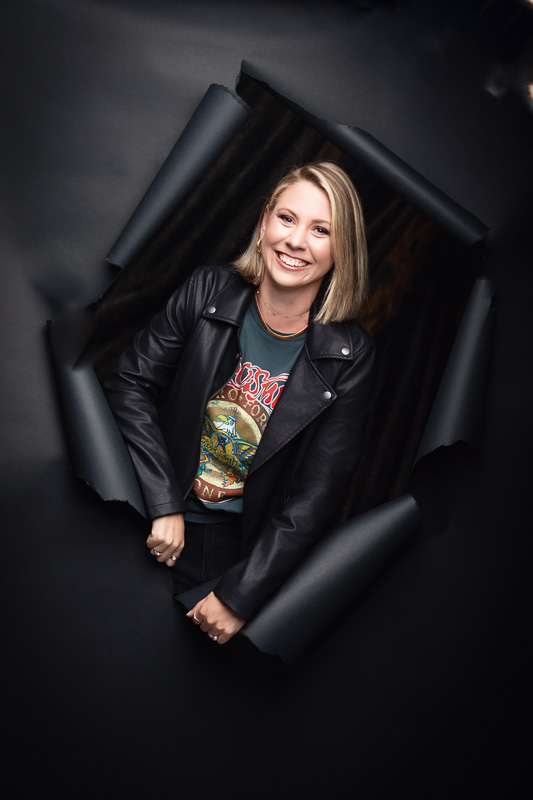 A person smiles while standing against a black background with torn paper edges. They are wearing a leather jacket over a graphic t-shirt. The surrounding torn paper gives a dynamic, emerging effect.