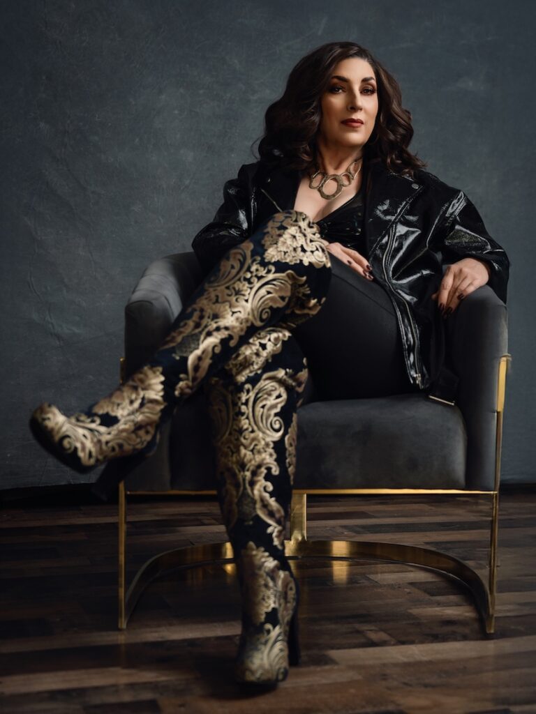A person with long dark hair sits confidently on a chair, wearing a shiny black jacket and ornate knee-high boots with intricate gold patterns. The background is a textured dark blue.