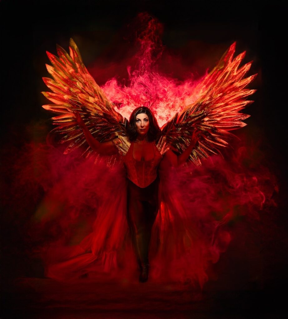 A person in a striking red and black outfit stands with large, fiery angel wings against a dark background. Flames surround the wings, creating a dramatic and mystical atmosphere.