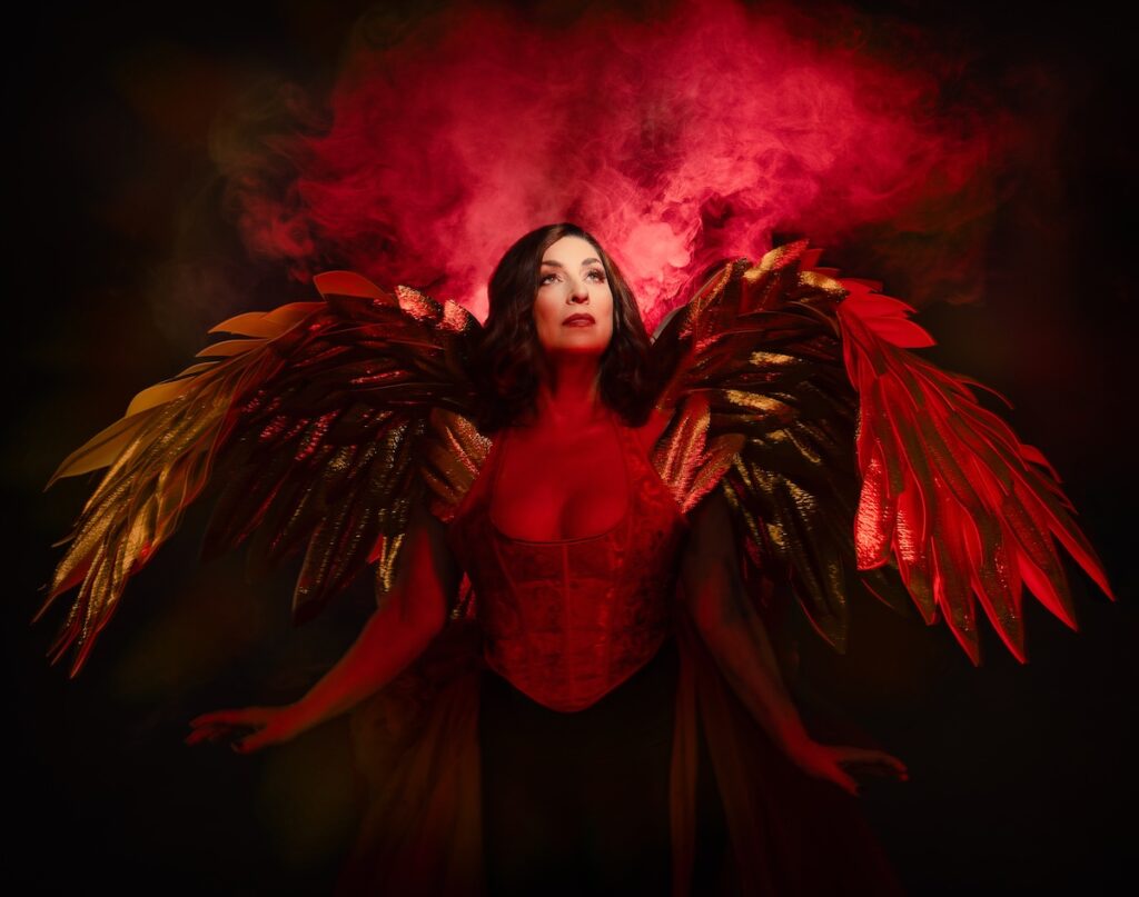 A woman in a fiery red and gold angelic outfit stands with large feathered wings. Red smoke billows behind her, creating a dramatic and mystical atmosphere. She gazes upward with a serene expression, illuminated by warm lighting.