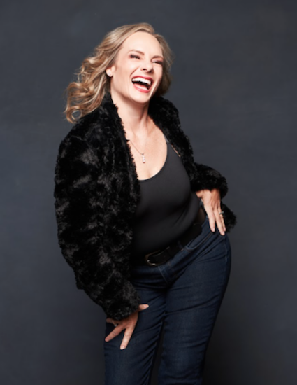 A person with long blond hair wearing a black fur jacket, black top, and jeans is laughing joyfully in front of a dark backdrop, striking a playful pose with hands on their hips.