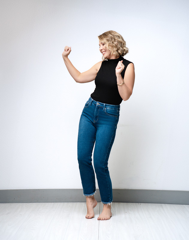 A woman with short blonde hair is joyfully dancing barefoot. She is wearing a black sleeveless top and blue jeans. The background is a simple white wall, and she appears relaxed and happy.
